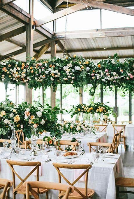 Rustic-chic wedding reception featuring long white-linen tables, honey-toned cross-back chairs, and lush greenery with roses in hanging floral installations.
