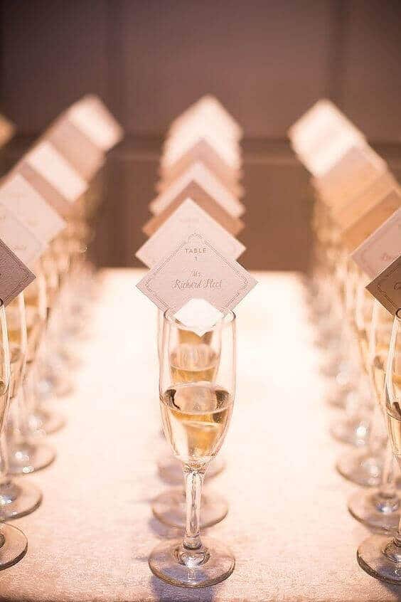 Elegant wedding seating display featuring champagne flutes with diamond-shaped name cards. Warm uplighting enhances the romantic atmosphere.
