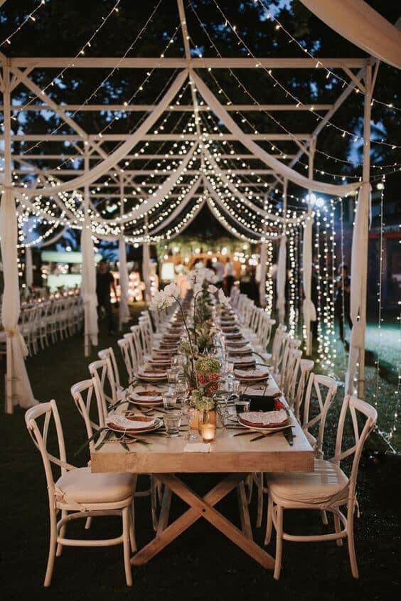 Elegant outdoor night wedding reception under a light-draped pavilion, featuring a long banquet table with rustic decor, warm fairy lights, and guests mingling.