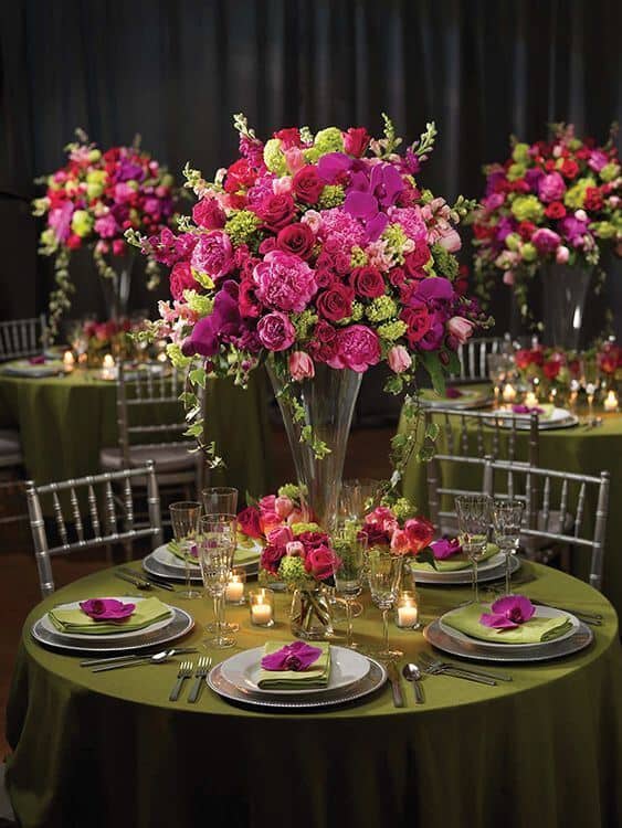 Luxurious wedding tablescape featuring a tall magenta and fuchsia floral centerpiece, olive green satin linens, silver chargers, and glowing votive candles.
