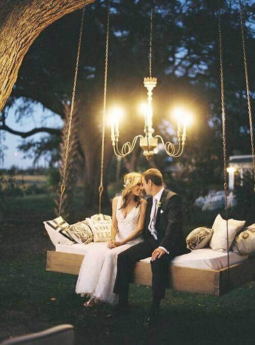 Bride and groom on a wooden swing-bed under a glowing chandelier, surrounded by pillows in an enchanting twilight garden setting.
