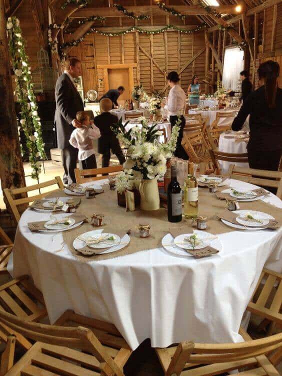 Round wedding table in a rustic barn, adorned with a burlap runner, white flowers, wine bottles, and kraft paper place cards, as guests mingle nearby.