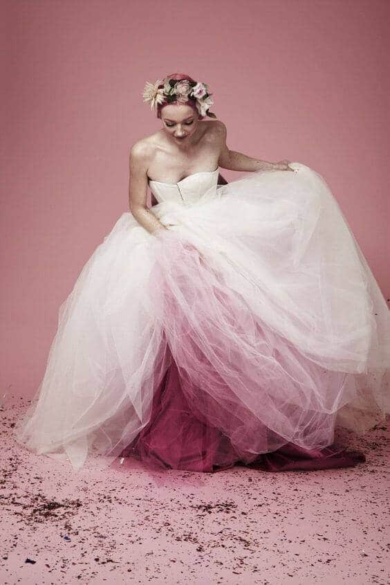 Bride in a voluminous white tulle gown lifting the skirt to reveal a berry-pink underlayer, adorned with a floral crown, against a blush backdrop.