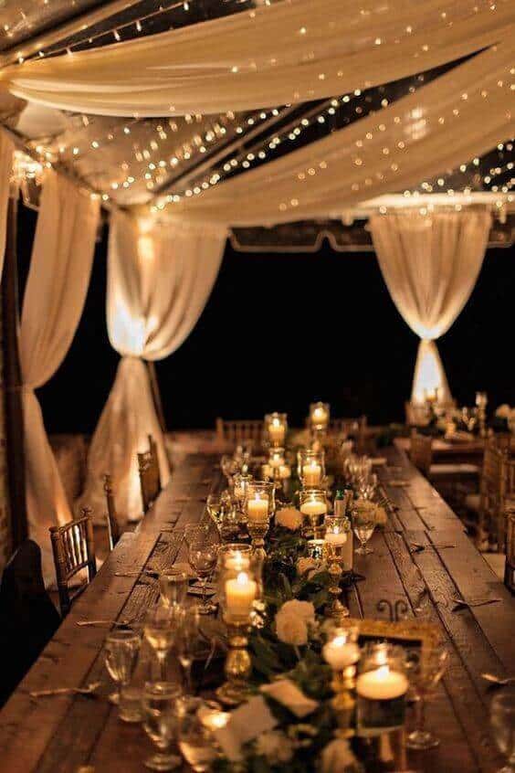 Candlelit tablescape under an ivory-draped tent, adorned with greenery and gold accents, creating a romantic evening ambiance for a wedding reception.