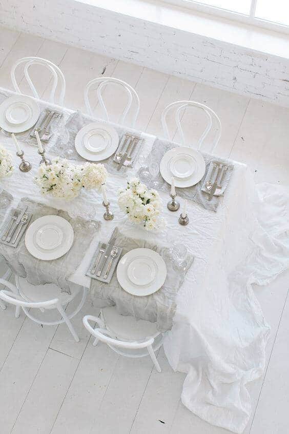 Elegant white-and-silver wedding reception table in a sunlit loft, featuring low floral arrangements, silver flatware, and unlit candles.
