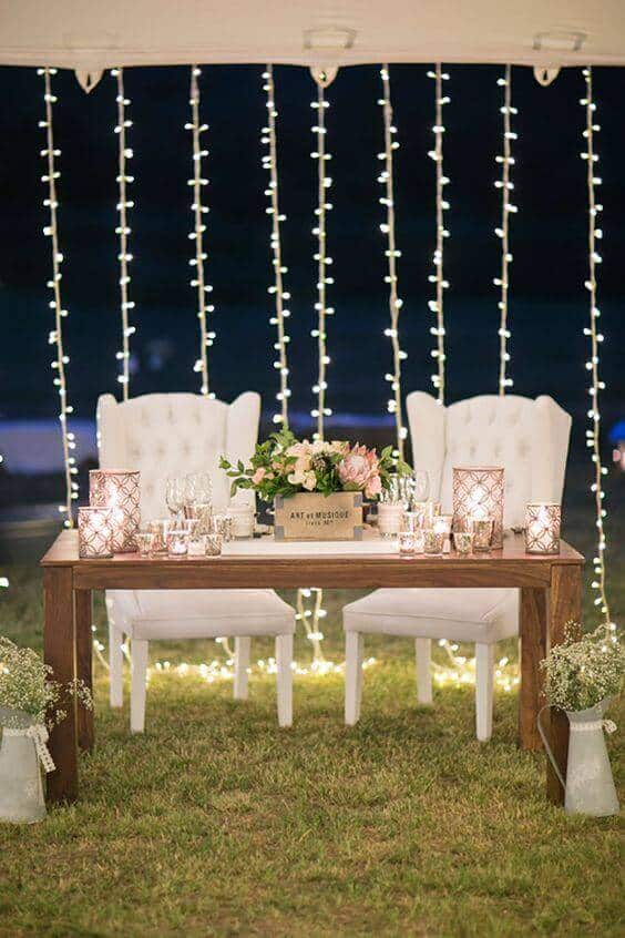 Outdoor evening sweetheart table with tufted ivory chairs, wooden table, fairy lights backdrop, candles, and blush floral centerpiece.