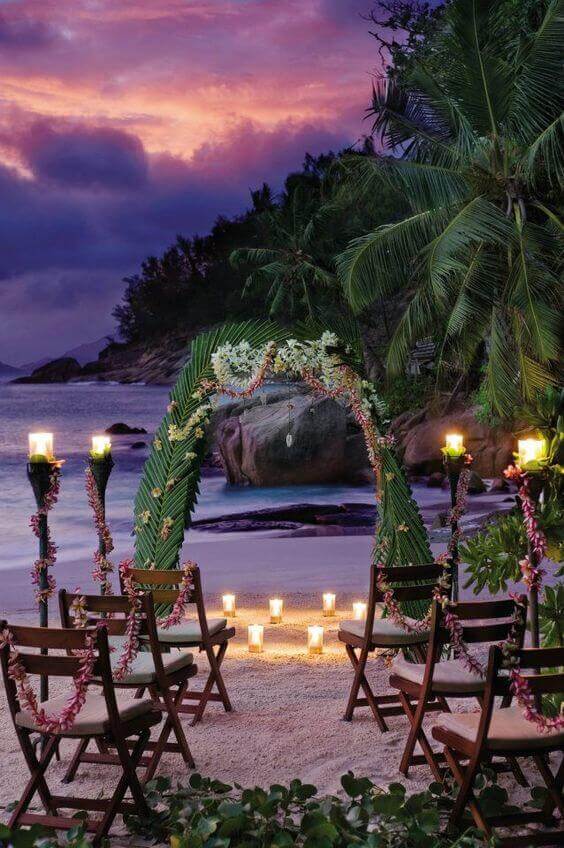 Romantic tropical beach night wedding setup with a palm-leaf arch, glowing votive candles, and tiki torches under a purple-pink sky.