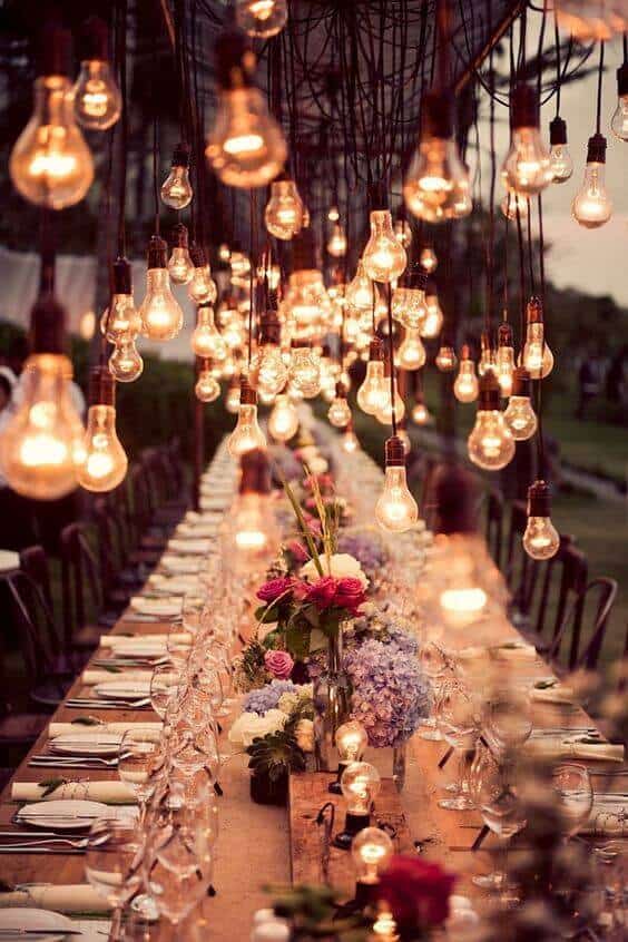 Outdoor wedding reception at dusk, featuring a rustic wooden banquet table adorned with lush hydrangea and rose centerpieces under a warm edison bulb canopy.