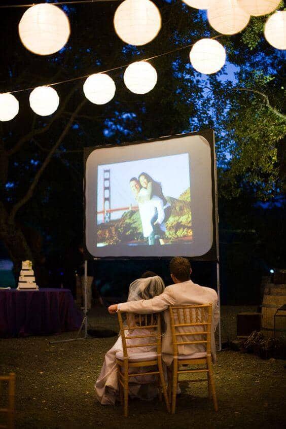 Bride and groom seated on Chiavari chairs, watching a slideshow under glowing paper lanterns at an outdoor evening wedding.