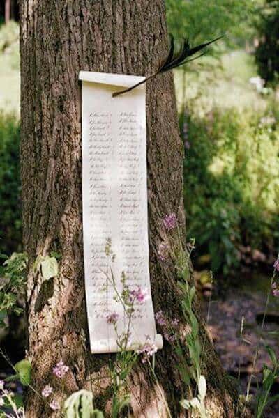 A cream calligraphy scroll hangs from a tree in a woodland, accented with a black feather quill, serving as a rustic wedding seating chart.