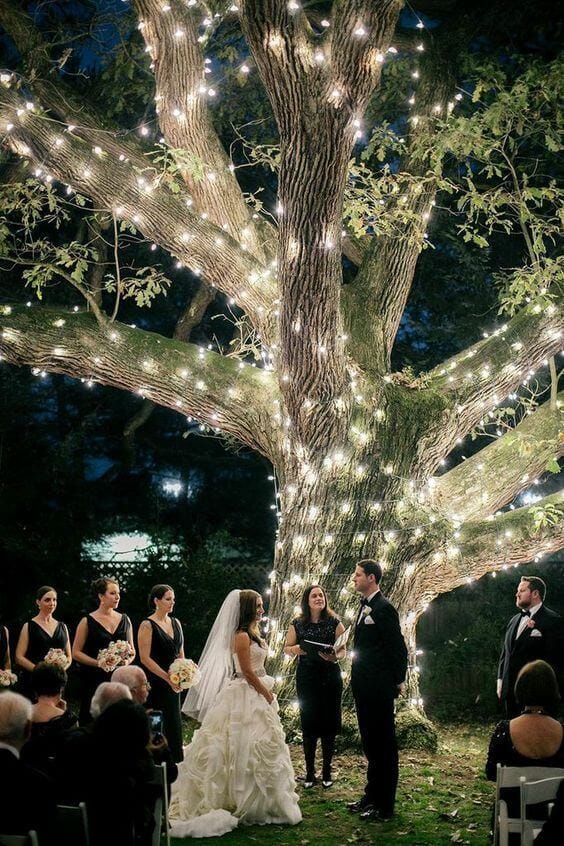 Nighttime outdoor wedding ceremony under a large oak tree adorned with warm white string lights, with the couple exchanging vows surrounded by guests.