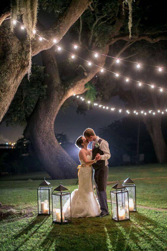 A newlywed couple embraces on a grassy lawn under oak trees adorned with warm-white string lights and glowing lanterns, creating a romantic night wedding scene.