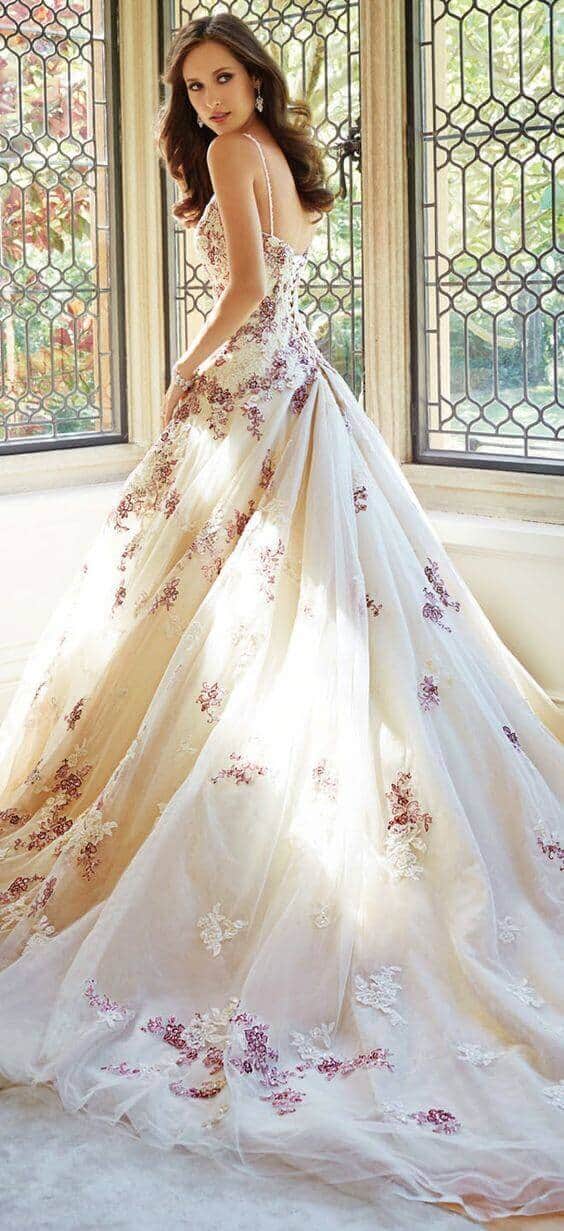 A bride in an ivory ballgown with a long train and floral lace appliqu&eacute;s stands gracefully in a sunlit alcove, framed by leaded-glass windows.