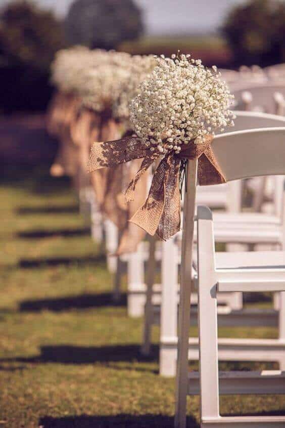Rows of white chairs on a sunlit lawn, each adorned with baby&rsquo;s breath bouquets and burlap bows, create a rustic wedding ambiance.