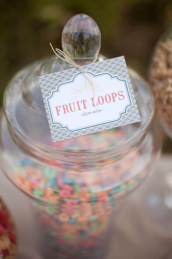 37 Morning Wedding Ideas filled with Sunshine color pallets 11 Clear apothecary jar filled with colorful Fruit Loops, labeled with a patterned tag, set at a whimsical wedding cereal bar.