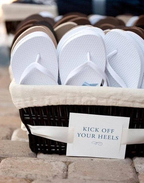 A dark wicker basket lined with ivory fabric holds white flip-flops, inviting guests to swap shoes at a beach wedding. A sign reads "kick off your heels. "