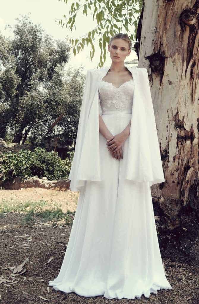 34 fancy wedding dresses and designs to make your big day sparkle 13 A serene bride in a flowing white gown with a lace bodice and shoulder-draped cape, standing by a eucalyptus tree in an outdoor garden setting.