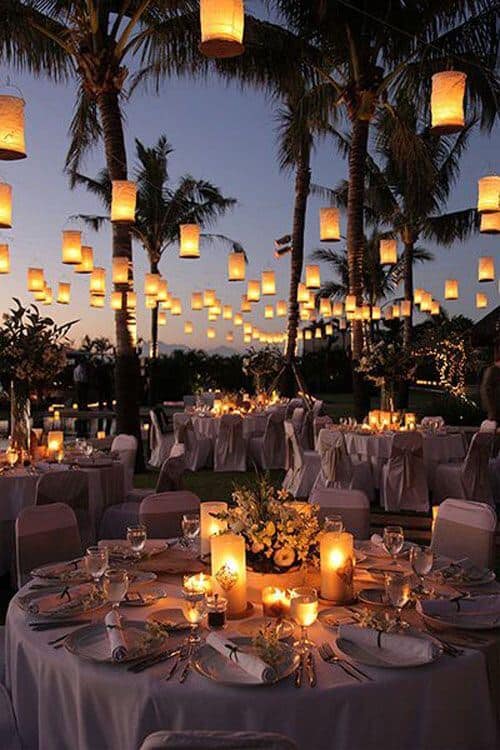 Outdoor beach wedding reception at dusk, featuring candlelit round tables, hanging lanterns, and palm trees creating a romantic atmosphere.