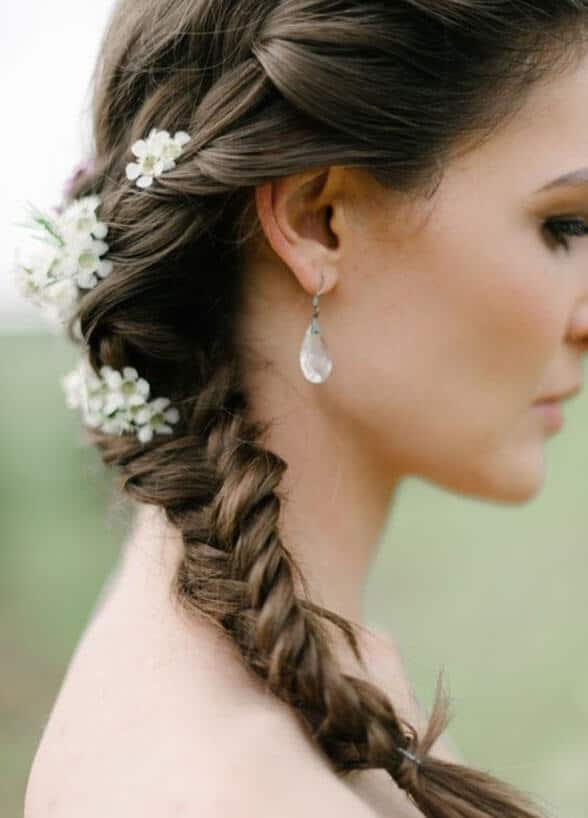 Bridal hairstyle featuring a textured side braid adorned with small white flowers, set against a soft green bokeh background.
