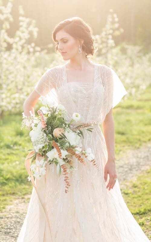 34 fancy wedding dresses and designs to make your big day sparkle 16 A bride in an ivory beaded gown with sheer flutter sleeves stands on a sunlit path in an orchard, holding a loose garden bouquet.