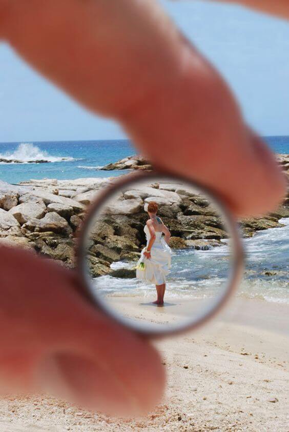 A hand holds a silver wedding band, framing a bride in an ivory dress at the shoreline, with turquoise waves behind her.