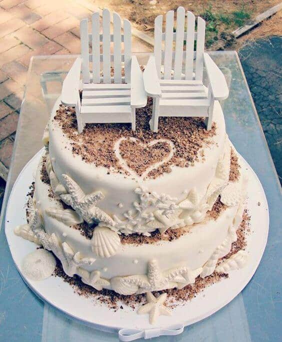 A two-tier beach wedding cake adorned with seashells and a heart motif, featuring miniature adirondack chairs, set outdoors.