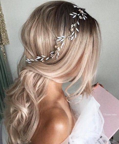 Side-profile of a bride with loose champagne-blonde waves, adorned with a silver leaf and crystal hair vine, draped in white tulle beside a blush-pink card.