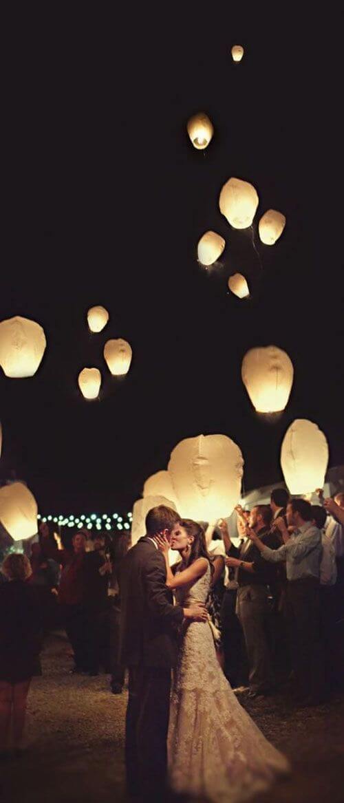 Newlyweds share a kiss under a canopy of warm amber sky lanterns, surrounded by guests celebrating a romantic beach wedding send-off.