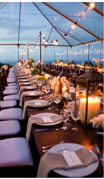 Elegant beach wedding reception scene under a clear tent at twilight, featuring wooden banquet tables, warm string lights, and white floral centerpieces.