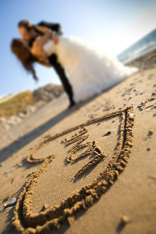 Foreground heart drawn in sand with "love," as a newlywed couple in white gown and dark suit share a dip-and-kiss by the ocean.