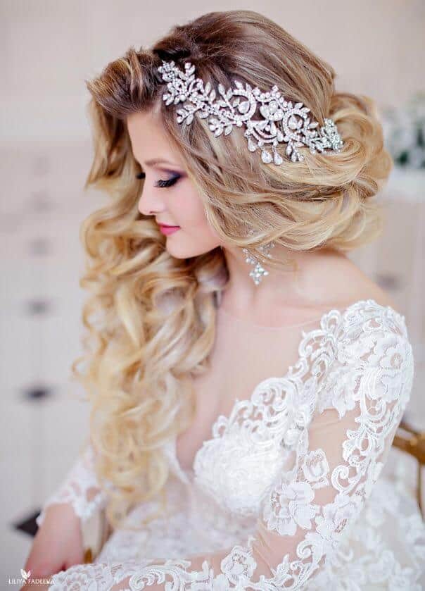 Bridal portrait showcasing cute wedding hairstyles with side-swept honey-blonde curls, adorned with a silver crystal headpiece and ivory lace gown.