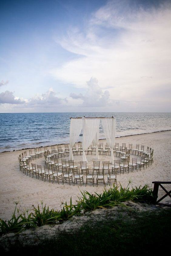 A serene beach wedding setup with light-wood chiavari chairs arranged in concentric circles around a white chiffon canopy, framed by a calm ocean.