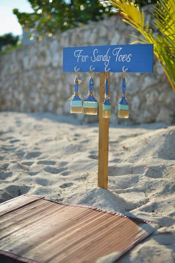Cobalt-blue sign reading "for sandy toes" with paintbrushes hanging below, set on a sandy beach with palm fronds in golden-hour light.