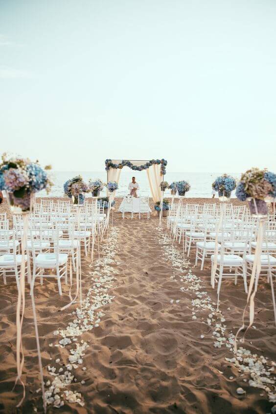Romantic beach wedding setup at golden hour: white chiavari chairs, petal-lined aisle, chiffon canopy, and blue hydrangea arrangements by the ocean.