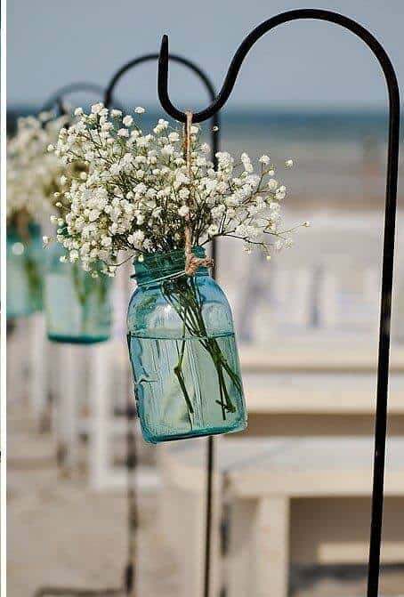 A sea-glass blue mason jar filled with baby's breath hangs by jute twine from a black shepherd's hook along a beach wedding aisle.
