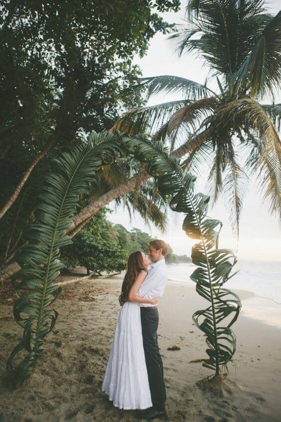 A couple kisses under a palm frond arch on a secluded beach at golden hour, surrounded by lush greenery and soft waves.