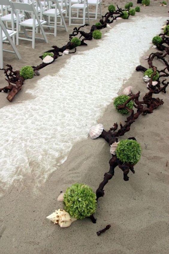 Coastal wedding ceremony aisle on soft sand, adorned with driftwood, seashells, and green pomanders beside white chairs.