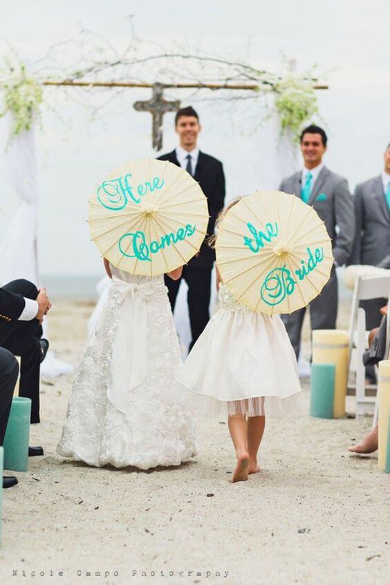 Two flower girls in ivory dresses walk barefoot down a sandy aisle, holding cream parasols with teal script, toward a driftwood arch.