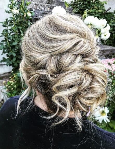 Textured low chignon hairstyle with honey-blonde and ash highlights, featuring loose tendrils, set against a garden backdrop with white blooms.