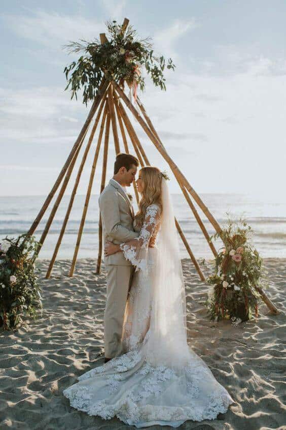 A bride in a lace gown and groom in a tan suit embrace beneath a teepee arch adorned with greenery and blush blooms on a sandy beach at sunset.