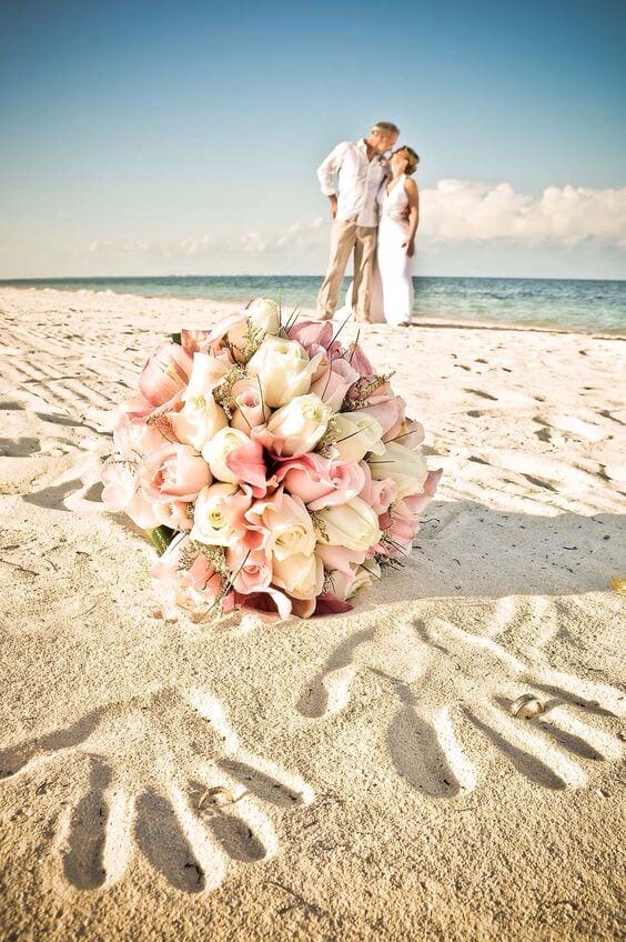 A romantic beach wedding scene featuring a pastel bouquet of blush calla lilies and ivory roses on sand, with a couple embracing nearby.