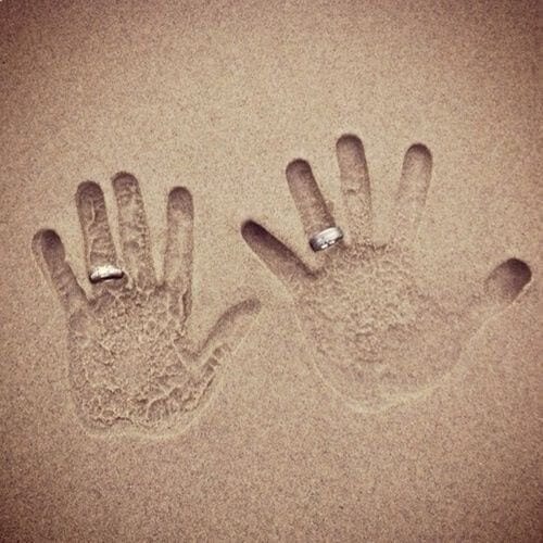 Two handprints in fine beach sand, each adorned with a wedding band, symbolize love and unity in a romantic coastal setting.