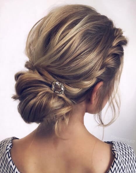 Romantic bridal hairstyle featuring a low chignon and side-swept braid, adorned with a crystal pin, set against a soft gray background.