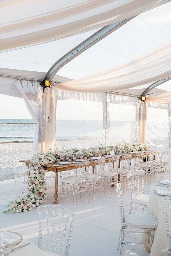 Beachfront wedding reception under a clear marquee featuring a long wooden table adorned with cascading white and blush florals, transparent chairs, and an ocean view.