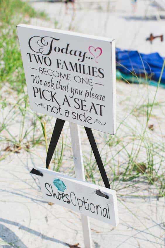 White wooden beach wedding sign reads "today… two families become one. We ask that you please pick a seat, not a side," with a smaller "shoes optional" plaque. Staked in sunlit sand with dune grass.