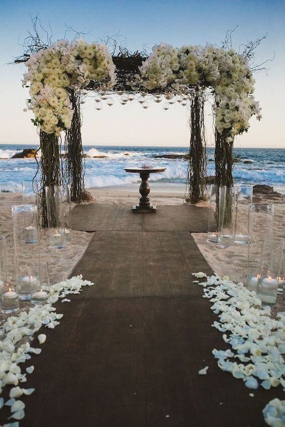 Romantic beach wedding setup featuring a floral arch with ivory roses, a wooden aisle on sand, and flickering candles in glass vases.