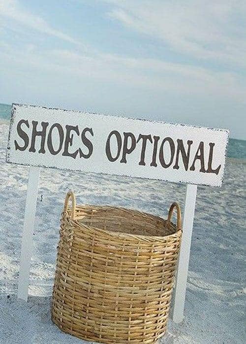 Whitewashed sign saying "shoes optional" above a wicker basket on sandy beach with teal ocean and blue sky, perfect for beach wedding ideas.