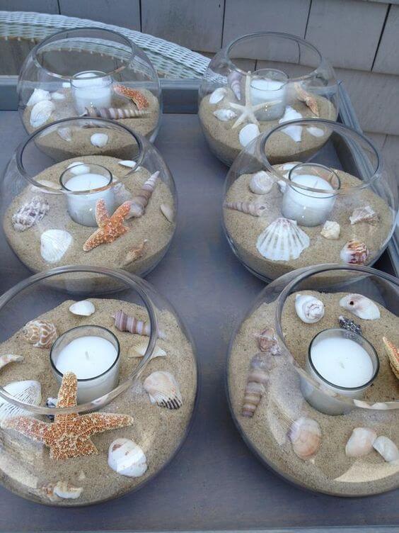 Coastal wedding centerpieces featuring glass bowls filled with sand, seashells, starfish, and tealight candles, arranged on a gray tray.