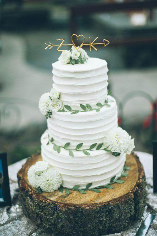Three-tier wedding cake with matte white buttercream, sage-green garland, white dahlias, and a gold wire z ♥ k topper on a wood-slice stand.