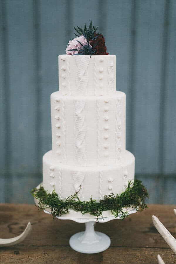 Rustic three-tier white wedding cake on a pedestal, adorned with cable-knit buttercream, moss, and a floral cluster of blush and burgundy blooms.